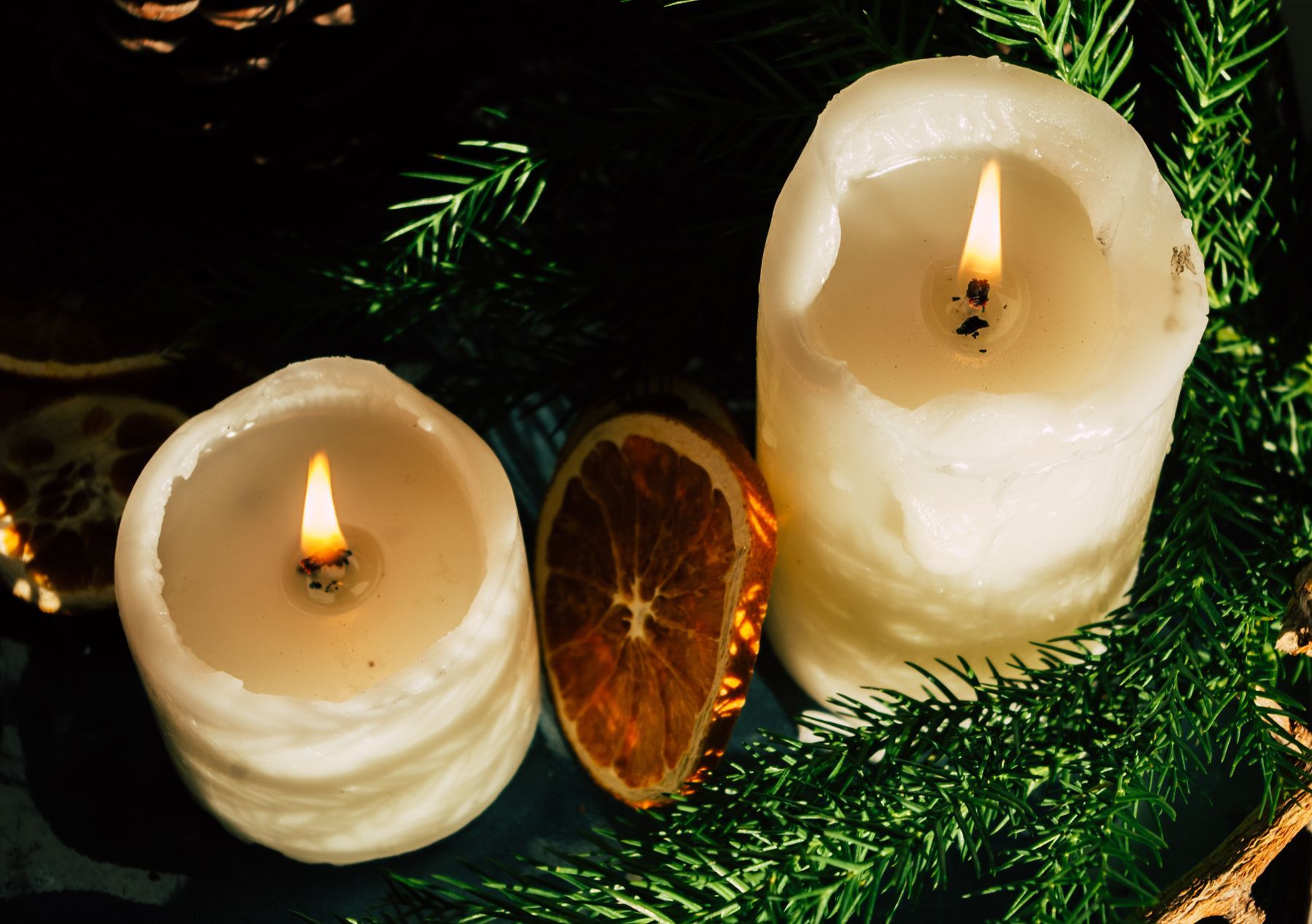 Two lit pillar candles with a slice of dried orange nestled in pine