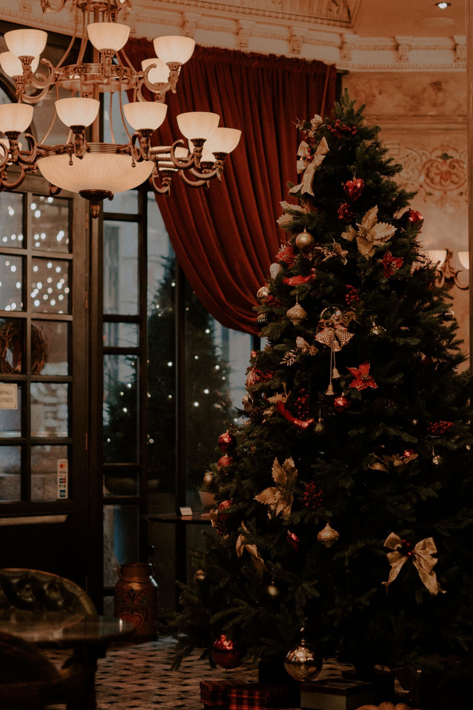 A richly dressed tree in a grand room with a chandelier and red velvet curtains