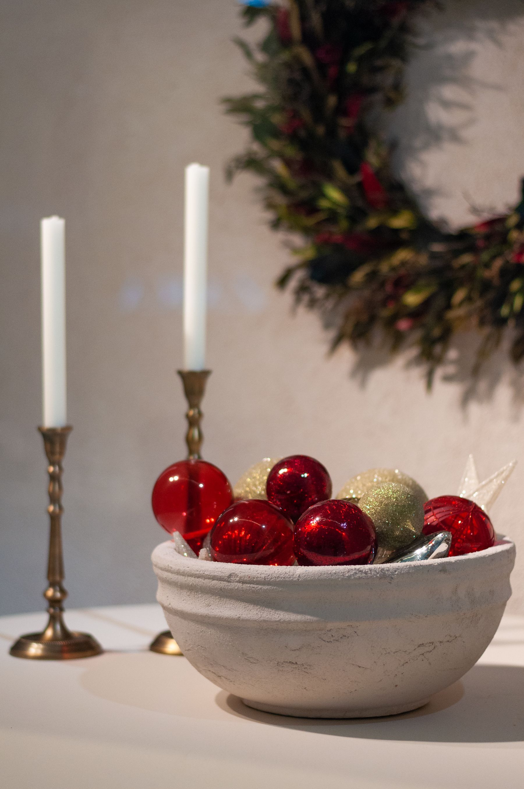 White taper candles in brass holders beside a stone bowl of glass baubles and a wreath