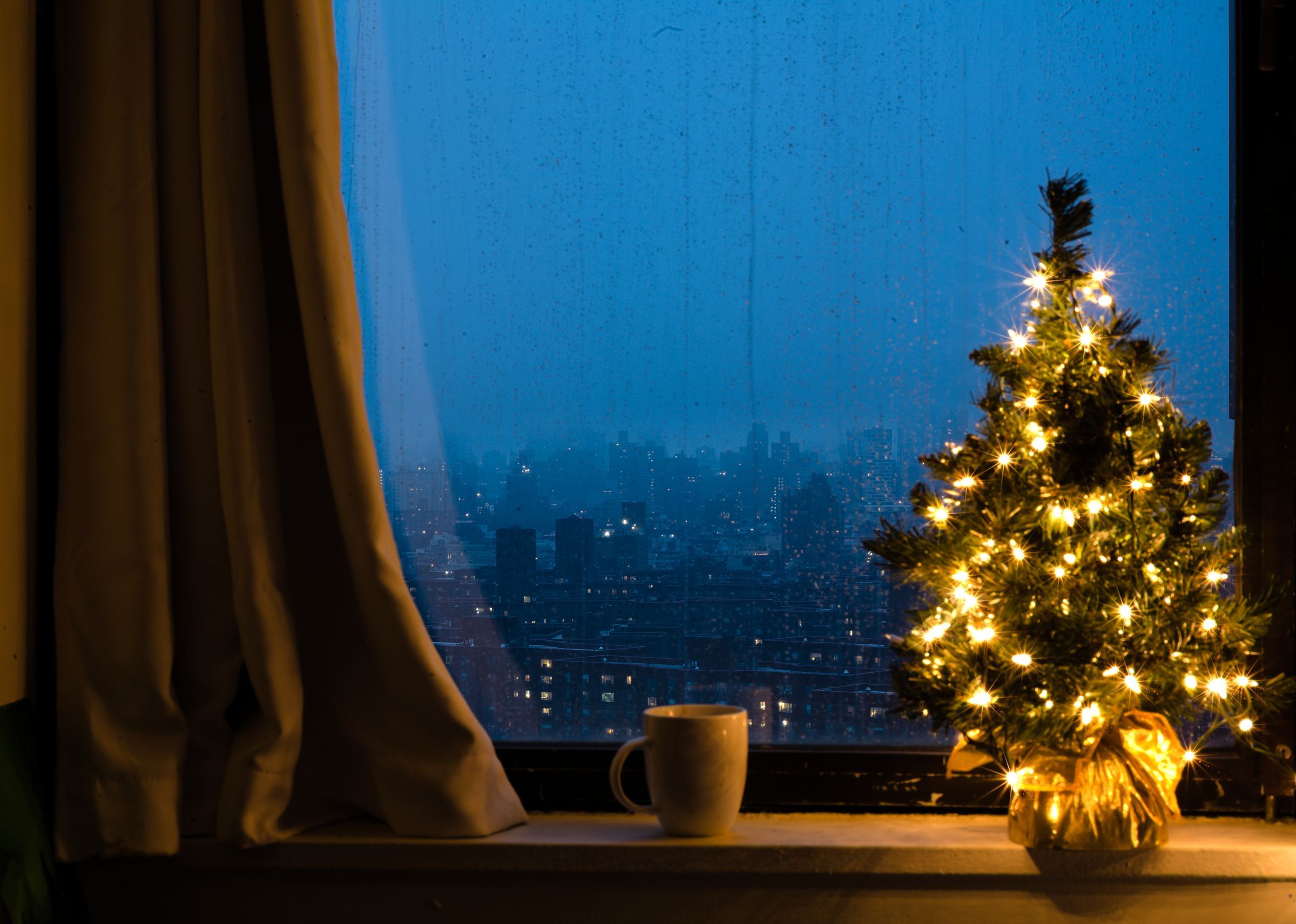 A small lit Christmas tree on a windowsill overlooking a rainy city at dusk